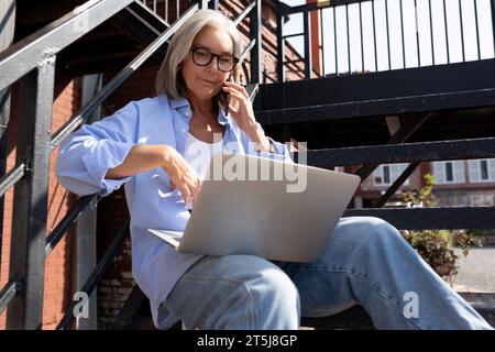 Reife grauhaarige Frau im Sommeroutfit arbeitet mit einem Laptop, während sie in der Stadt auf der Straße sitzt Stockfoto