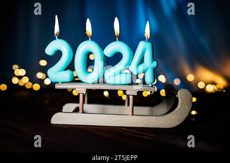 Feiertagshintergrund Happy New Year 2024 von blauen Kerzen beleuchtet auf einem Schlitten auf einem blauen Hintergrund mit Weihnachtslichtern. Neujahrsfeiertag, ganz in der Nähe Stockfoto