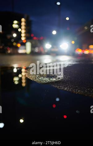 Unscharfe Sicht auf Autos, die nachts auf Straßenhöhe vorbeifahren, mit Reflexion in der Pfütze Stockfoto