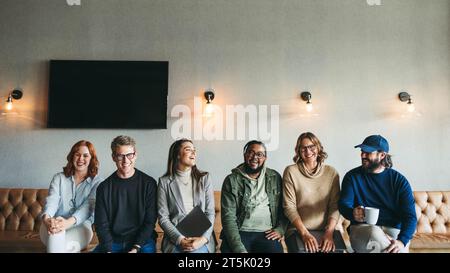 Ein vielfältiges Team von Geschäftsleuten sitzt zusammen in einem Startup-Büro. Die Kolleginnen und Kollegen, Männer und Frauen, lächeln glücklich und spiegeln eine Zusammenarbeit und Erfolg wider Stockfoto