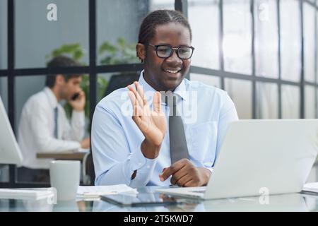 Ein Mann am Arbeitsplatz an einem Tisch vor einem Computer begrüßt Kollegen per Videokommunikation Stockfoto