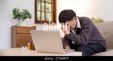 Asiatischer reifer Erwachsener, der an Fieber leidet, seinen Arzt anruft und seinen Kopf berührt, auf der Couch vor dem Laptop im Wohnzimmer sitzt Stockfoto