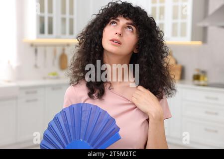 Junge Frau winkt mit blauem Handventilator, um sich in der Küche zu kühlen Stockfoto