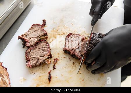 Grill Restaurant Küche. Koch in schwarzen Kochhandschuhen mit Messer zum Schneiden von geräuchertem Schweinefleisch Stockfoto