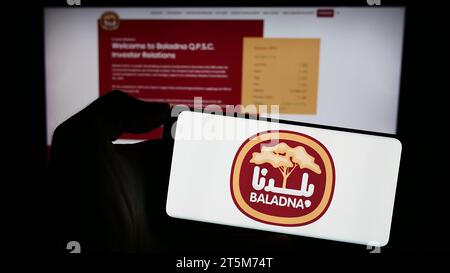 Person mit Handy mit Logo des katarischen Molkerei- und Lebensmittelunternehmens Baladna vor der Business-Webseite. Schwerpunkt auf dem Display des Telefons. Stockfoto