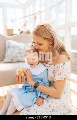 Vertikale Aufnahme der lächelnden jungen Mutter und der entzückenden Tochter des Kleinkindes im Kleid, die mit kleinen gelben Entlein im Sommerhaus mit Pavillon an sonnigen Tagen spielt. Stockfoto