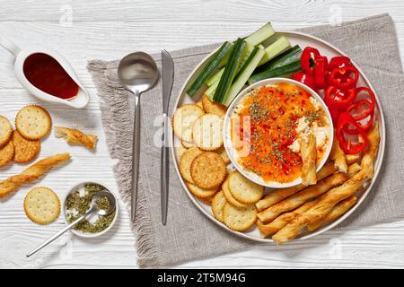 Hot Pepper Jelly Cheese Dip in Bowl serviert mit Crackern, salzigem grissini oder knackigen italienischen Brotstangen, frischem Pfeffer und Gurke auf Platte auf weißem W Stockfoto