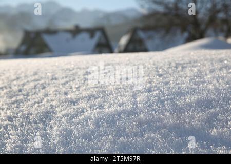 Purer, unberührter Schnee. Nahaufnahme. Auf der welligen Oberfläche funkeln Schneeflocken in der Sonne. Dorfhäuser im Hintergrund Stockfoto