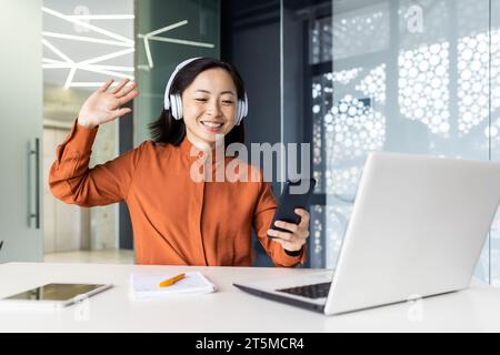 Junge Frau im Büro am Arbeitsplatz, die entspannende, beruhigende Musik hört, asiatische Geschäftsfrau in Kopfhörern mit App am Telefon, sitzt mit Laptop am Tisch, trägt Kopfhörer. Stockfoto