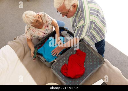 Glückliches kaukasisches Seniorenpaar, das Pässe in den Koffer packt, in einem sonnigen Schlafzimmer zu Hause. Pensionierung, Zusammengehörigkeit, häusliches Leben und Lebensstil der Senioren, Stockfoto