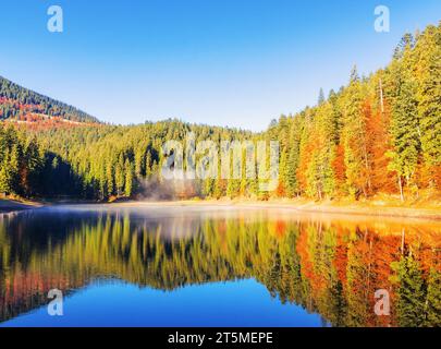Herbstlandschaft mit See. Sonniger Morgen mit blauem Himmel und Nebel. Bewaldete Hügel spiegeln sich auf der Wasseroberfläche. Stockfoto