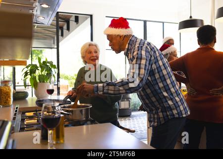 Glückliche, vielfältige Freunde, die sich mit weihnachtsmann-Hut in der sonnigen Küche zuhause zubereiten. Ruhestand, Freundschaft, weihnachten, Feiern kochen, essen, Senior Stockfoto