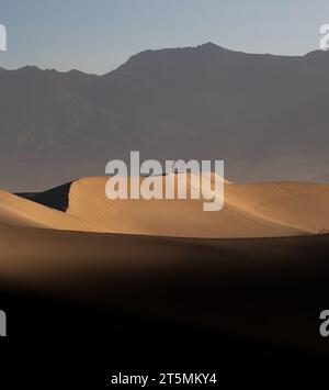 Death Valley, USA. Mesquite flacht am frühen Morgen Sanddünen ab. Ein Mann, der allein auf der Düne steht. Hoher Berg im Hintergrund. Klarer Himmel Stockfoto