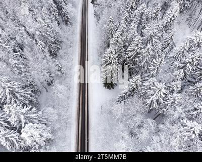 Drohnenblick auf eine Asphaltstraße inmitten eines wunderschönen schneebedeckten Waldes. Stockfoto