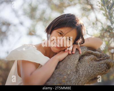 Schöne asiatische junge Frau in weißem Kleid draußen in der Nähe von Olivenbäumen. Genießen Sie frische Luft und nehmen Sie an Outdoor-Aktivitäten Teil. Friluftsliv Konzept bedeutet, so viel Zeit wie möglich im Freien zu verbringen Stockfoto