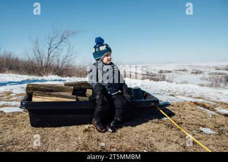 Seitenansicht eines kleinen Jungen, der auf einem Schlitten voller Feuerholz in Schnee sitzt Stockfoto