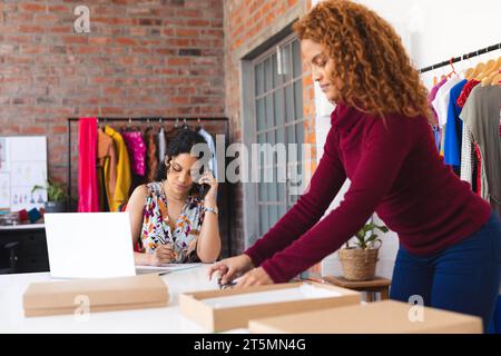 Zwei zweirassige Modedesignerinnen packen Pakete mit Smartphone und Laptop in einem sonnigen Studio Stockfoto