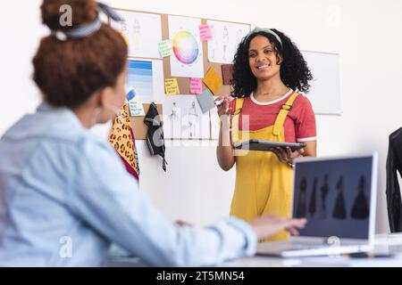 Zwei zweirassige Modedesignerinnen diskutieren über Tablet und Laptop im sonnigen Studio Stockfoto