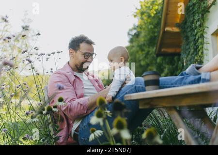 Mann sitzt im Garten mit Füßen auf dem Tisch und spielt mit seinem kleinen Sohn. Vater hat Bindungsmoment mit seinem kleinen Kind. Stockfoto