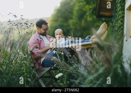 Mann sitzt im Garten mit Füßen auf dem Tisch und spielt mit seinem kleinen Sohn. Vater hat Bindungsmoment mit seinem kleinen Kind. Baby spielt mit Blume. Stockfoto