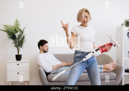 Frau tanzt mit schnurlosem Vakuum, während Mann mit Laptop Stockfoto