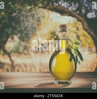 Eine Flasche handgefertigtes Olivenöl steht auf einem Holztisch, hinter einem Olivengarten an einem etwas Bokeh, sonnigen Tag. Bild im Vintage-Stil. Stockfoto