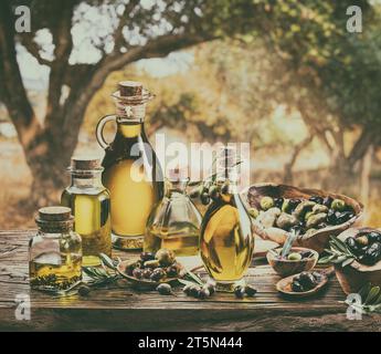 Eine Flasche handgefertigtes Olivenöl steht auf einem Holztisch, hinter einem Olivengarten an einem etwas Bokeh, sonnigen Tag. Bild im Vintage-Stil. Stockfoto