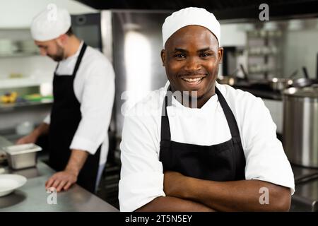 Porträt eines glücklichen afroamerikanischen Küchenchefs, der mit überkreuzten Armen in der Restaurantküche steht Stockfoto