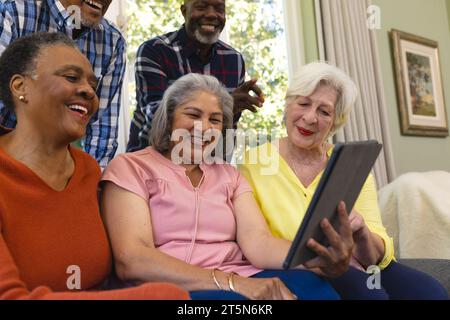 Glückliche, vielfältige Gruppe von älteren Freunden, die Videoanrufe machen und im sonnigen Wohnzimmer lachen Stockfoto