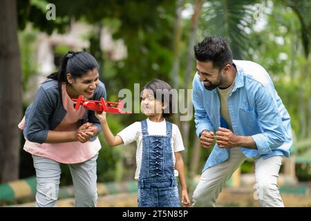 Tracking-Aufnahme von aufgeregten indischen Eltern, die mit einem Mädchen laufen, während das Kind mit einem Spielzeug-Flugzeug im Park spielt - Konzept der aktiven Erziehung, Familie Stockfoto