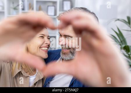 Älteres Ehepaar, Mann und Frau, die das Herz zeigen, mit den Händen vor die Kamera, verliebter Mann und Frau sitzen auf dem Sofa im Wohnzimmer, lächeln und umarmen. Stockfoto