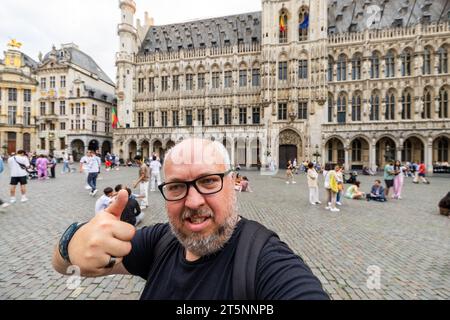 Inmitten der berühmten Wahrzeichen und lebhaften Straßen Brüssels hält ein männlicher Tourist mit markanten grauen, kurzen Haaren, Bart und Brille, der 40 Jahre alt ist, an, um den Moment zu ergreifen. Mit seinem Smartphone in der Hand macht er ein jubelndes Selfie, das einen Daumen nach oben zeigt, ein Symbol für seine Wertschätzung und Freude an der Erkundung des Charmes der Stadt. Fröhliches Selfie für Touristen: Brüssels Herz erkunden. Hochwertige Fotos Stockfoto