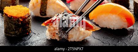 Sushi mit Stäbchen Panorama. Unagi-Sushi, Nigiri mit Aal und anderes Sushi und Brötchen auf schwarzem Hintergrund. Japanisches Restaurant Stockfoto