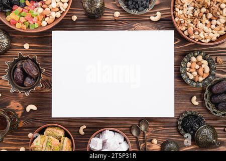 Leeres weißes Papier, umgeben von gemischten getrockneten Früchten, Datteln aus Lukum, Baklava, Nüssen, Holzschreibtisch ramadan Stockfoto