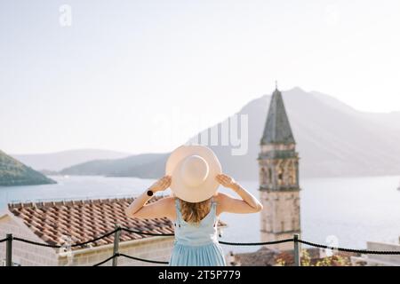 Das Mädchen steht auf der Aussichtsplattform in der Nähe des Glockenturms und hält ihren Hut mit den Händen. Perast, Montenegro. Rückansicht Stockfoto