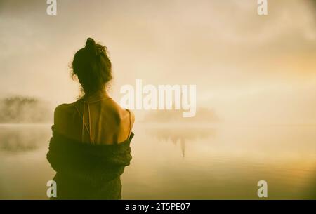 Frau im grauen Mantel, die einen Sonnenaufgang im Nebel am Ufer des Bleder Sees, Slowenien V, beobachtet Stockfoto