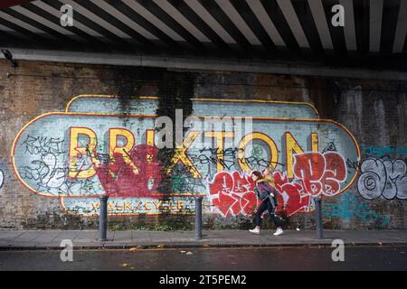 Eine Frau gibt einem Kind einen Huckepack unter einer Eisenbahnbrücke und vorbei an einem großen Wandbild von Brixton in Brixton South London, am 4. November 2023 in London, England. Stockfoto