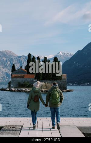 Mann und Mädchen laufen am Pier entlang und schauen auf die Berge hinter der Insel St. George. Montenegro. Rückansicht Stockfoto