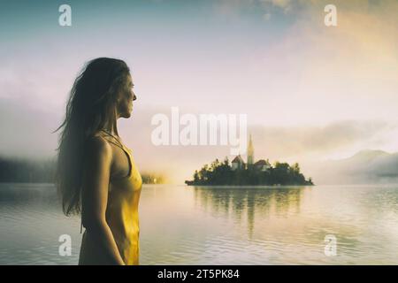 Frau in grauem Mantel und gelbem Kleid, die einen Sonnenaufgang im Nebel am Ufer des Bleder Sees, Slowenien VI., beobachtet Stockfoto