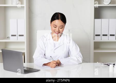 Lächelnde Asiatische Ärztin Schreibt Notizen, Während Sie Am Schreibtisch In Der Klinik Sitzt Stockfoto