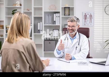 Rückansicht, erfahrener reifer Arzt berät lächelnde Patientin, grauhaariger Mann, der in der Klinik arbeitet, Laptop benutzt. Stockfoto