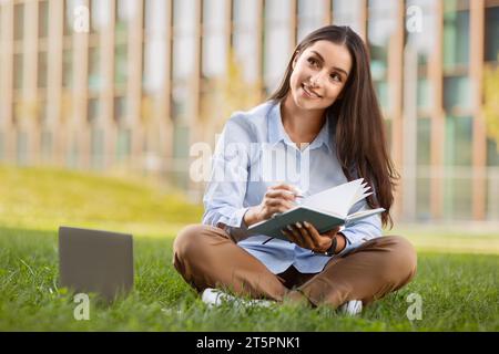 Glückliche nachdenkliche hübsche tausendjährige kaukasische Dame mag Arbeit mit Laptop, Schreiben in Notizbuch Stockfoto
