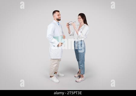 Junger männlicher Arzt in Augenheilkunde berät Frau, grauer Hintergrund Stockfoto