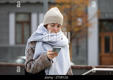 Mädchen in Strickmütze und Schal, das mit Smartphone in den Händen auf der Herbststraße läuft Stockfoto