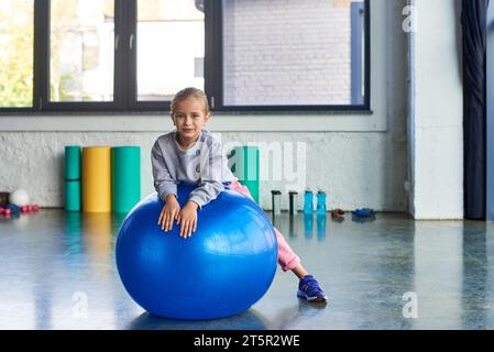 Hübsches kleines Mädchen in Sportbekleidung, das auf Fitnessball liegt und leicht in die Kamera lächelt, Kindersport Stockfoto