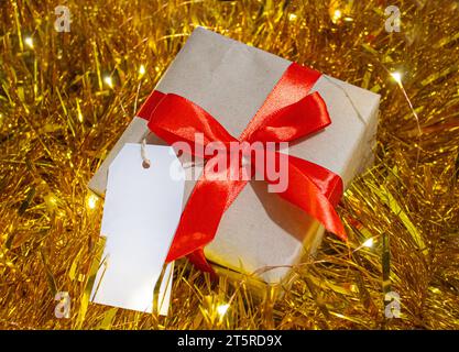 Geschenkbox verpackt mit Kraftpapier und roter Schleife im goldenen Weihnachtsgirlande Hintergrund mit weißem Tag für Text. Winterferien Einkaufen und Feiern c Stockfoto