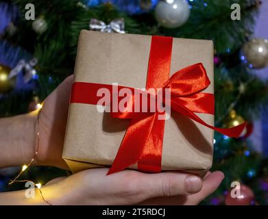 Mann, der in Händen schöne Weihnachtsgeschenkbox in Kraftpapier und rotem Band auf dem Hintergrund von Weihnachtsbaum und Lichtern hält. Präsentiert Vorbereitung auf Stockfoto
