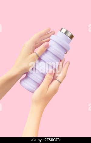 Frauenhände halten zusammenklappbare wiederverwendbare Wasserflasche in Flieder auf rosa Hintergrund. Nachhaltiger Lebensstil Stockfoto