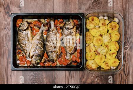 Gebackene Brasse mit Gemüse auf einem Backblech steht mit gebackenen Kartoffeln in einer Schüssel auf einem Holztisch. Draufsicht Stockfoto