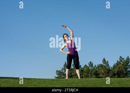 Sportliche junge Frau, die sich vor dem Fitnesstraining im Park warm-up macht, blauer Himmel Hintergrund. Eine schöne Frau praktiziert Yoga, Pilates, barfuß auf Stockfoto